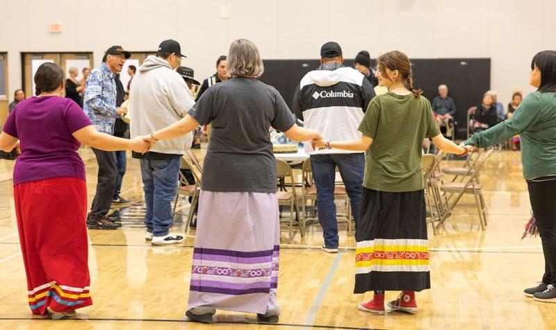Four women wearing ribbon skirts hold hands as they dance around Indigenous drummers