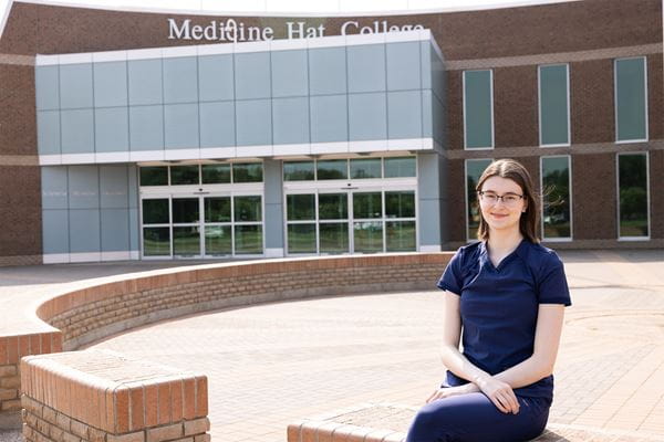 Health Care Aide Alumni Student, Emma Mourton, sitting in front of the main entrance of Medicine Hat College. 