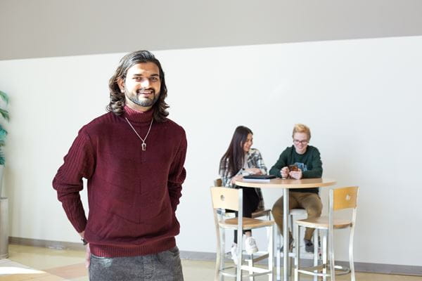Student standing in Centennial Hall with two students sitting at table in the background