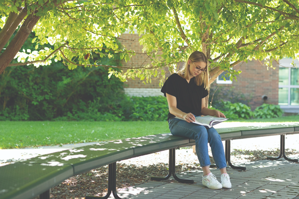 A student sitting outside on a bench under a tree looking down at her textbook.