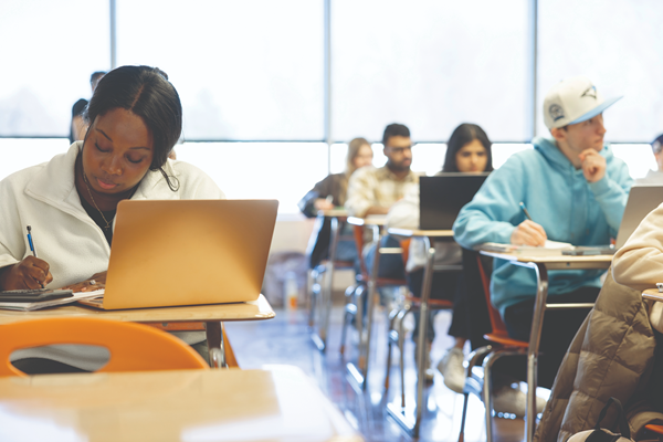 Students learning in a classroom, with one student taking notes with her laptop open