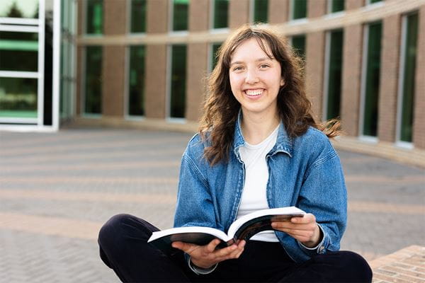Student sitting at front of MHC with a book open, smiling at the camera. 