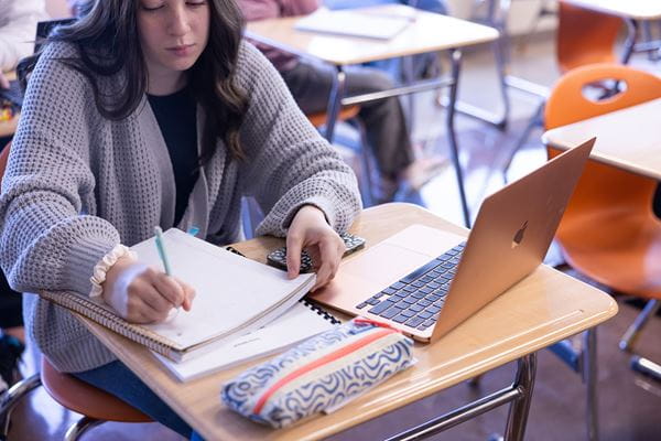 Person at desk with an open laptop while writing notes on notebook.