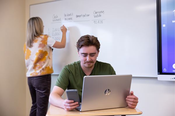 Student working on laptop with calculator as the instructor writes on the whiteboard in the background