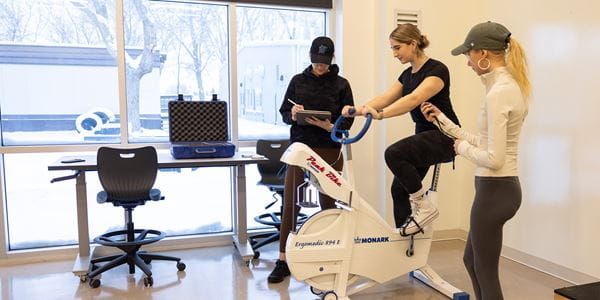 Student riding a fitness bike as two classmates complete an assessment
