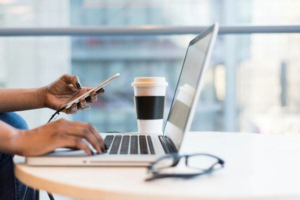 Close up of someone working on a laptop, holding a phone with a travel coffee cup and a pair of glasses. 