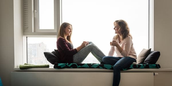 Mother and daughter sit on window ledge enjoying coffee