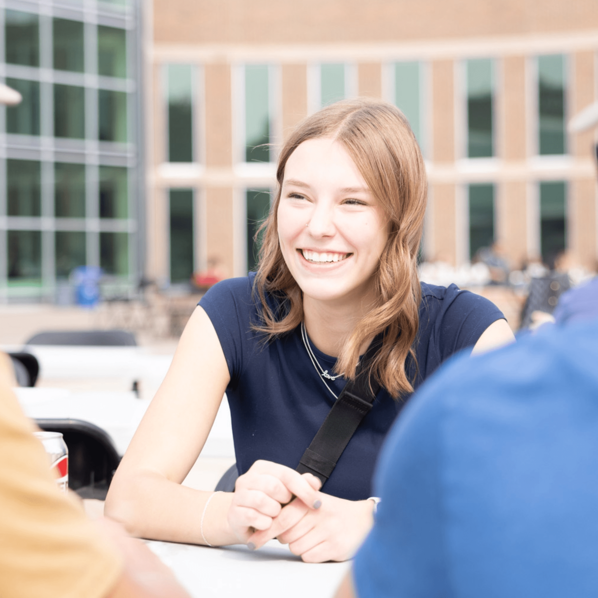 New students sitting at table, girl smiling and looking at friend, with MHC campus behind. 