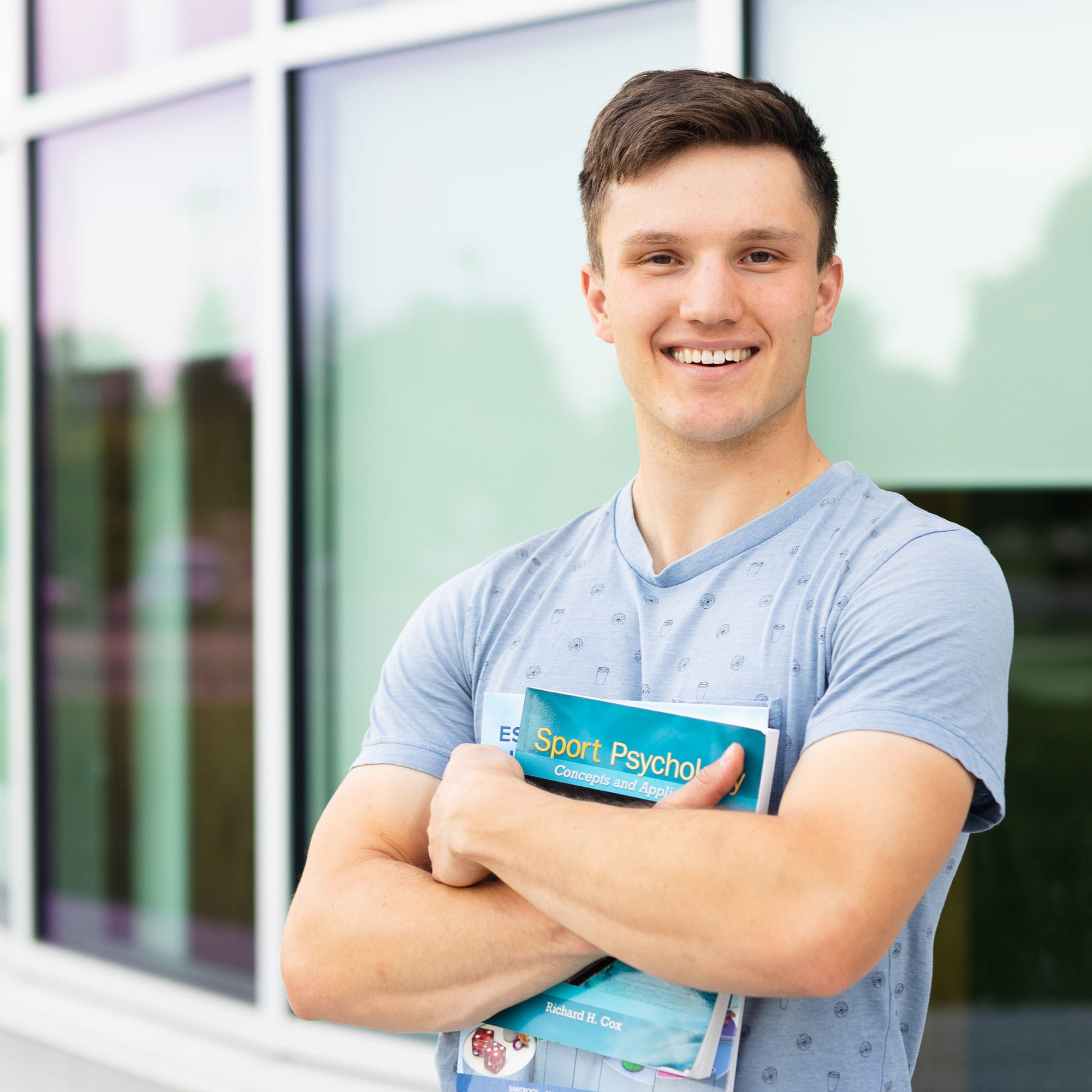 Student standing in front of windows holding books with arms crossed. 