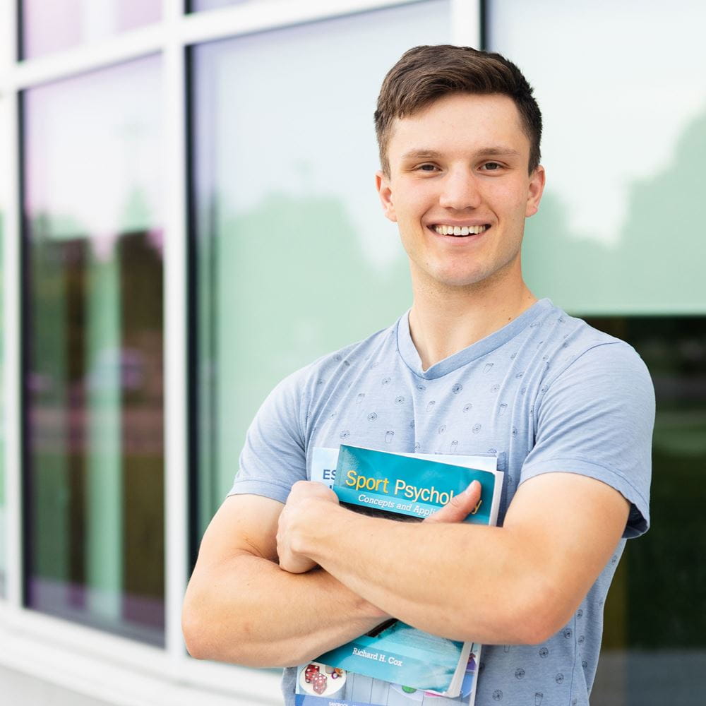 Student standing in front of windows holding books with arms crossed.