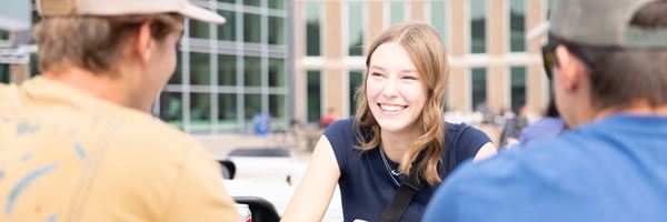 New students sitting at table, girl smiling and looking at friend, with MHC campus behind.