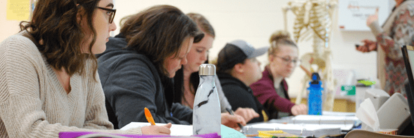 Students taking notes at a desk in a classroom. Anatomy skeleton behind students and instructor teaching in front of them. 