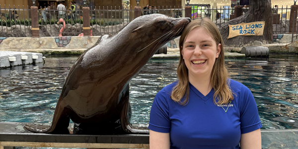 MHC Service Dog & Canine Studies alumni, Megan Jingle smiling at camera while sea lion kisses her head.