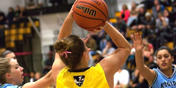 Close up of a female basketball player about to through a basketball. Opposing teammates around her with crowd behind on bleachers.
