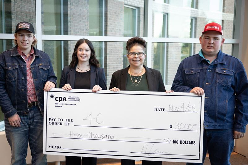 Two male students in denim jackets and ballcaps hold either end of a large cheque with two brown-haired women holding it in the middle.