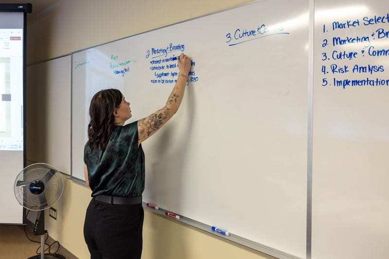 A female student with tattoos writes on a whiteboard under the heading Marketing and Branding.