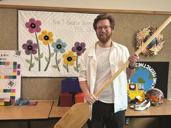 A male student with brown hair and facial hair holds a canoe he crafted for a project in the Bachelor of Education program