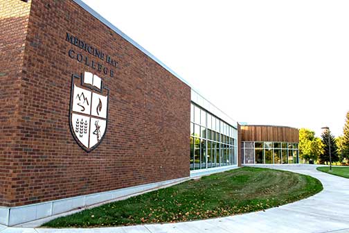 A brown brick building with glass windows, green grass, a sidewalk, and the Medicine Hat College logo