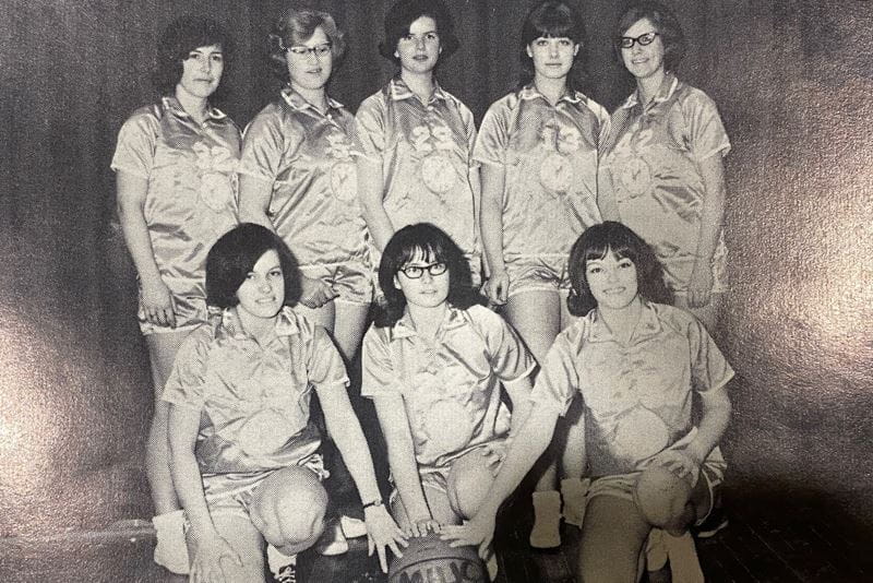 A black and white photo from 1965 with eight women posing with a basketball with MHC written on it.