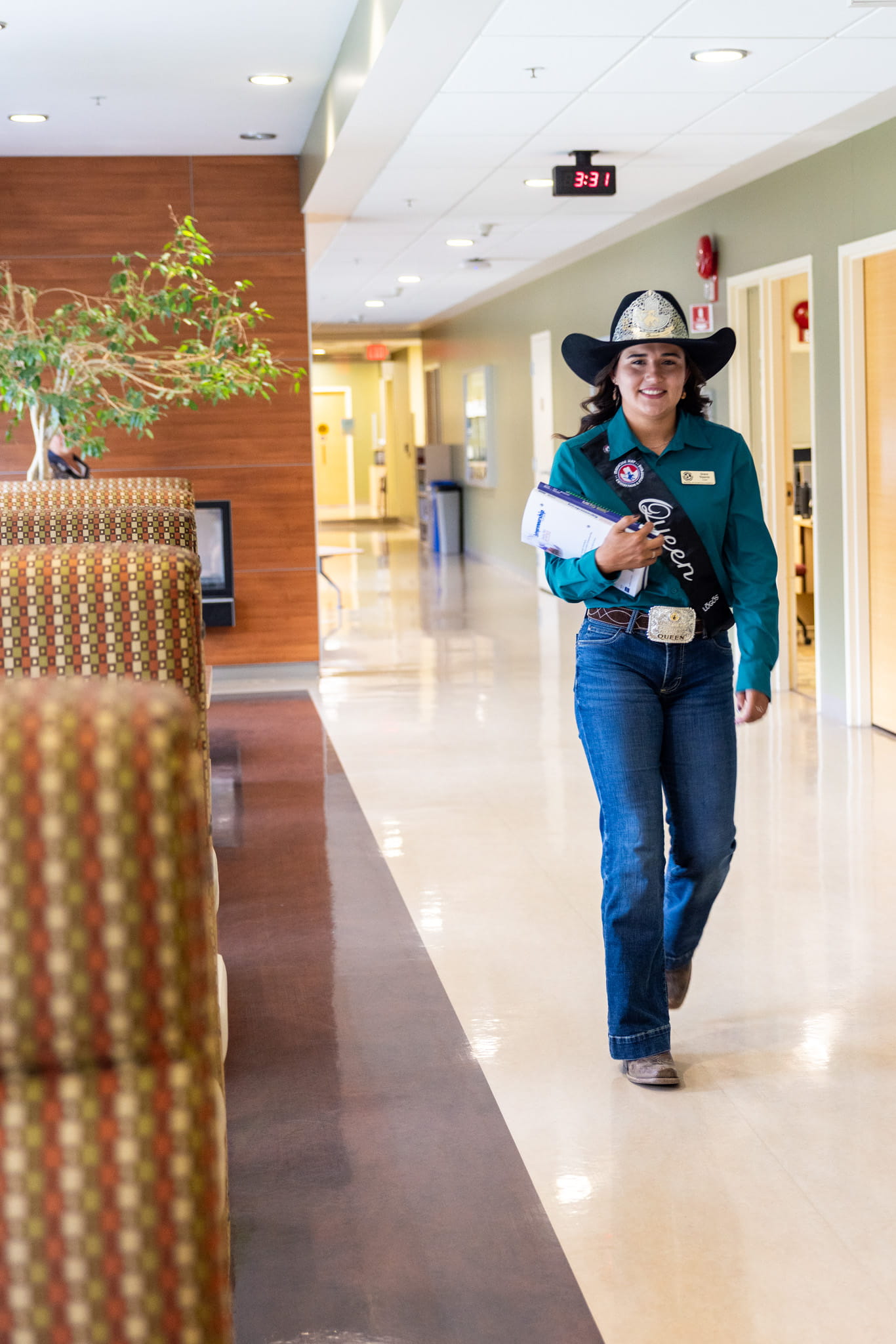 Woman with cowboy hat and green shirt walks down hall with textbooks in hand.