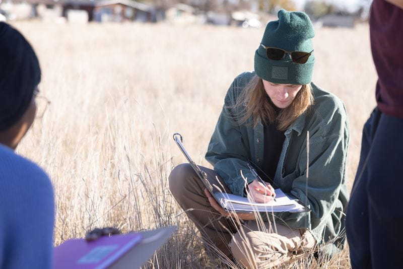 A male student wearing a touque and sunglasses documents his findings in a binder. 