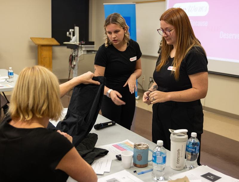 Two female business students stand in front of the judges as they assess the prototype they created for Venture Design Studio, a pair of pants for women in the trades