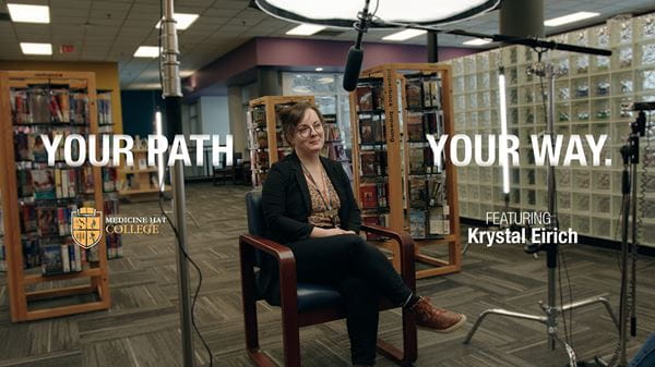 Krystal Eirich, MHC alumni, seated in middle of library with camera equipment around her and graphic text saying, 