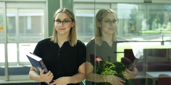Student holding books, leaning against a glass window