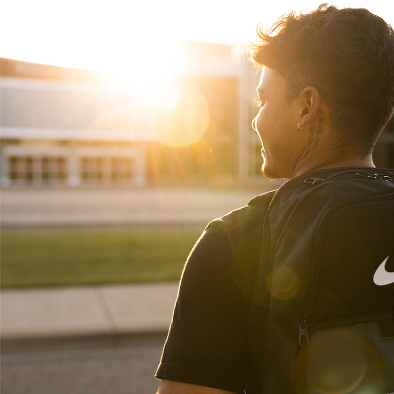 Student in front of MHC, wearing a backpack, with sun shining. 